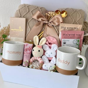 May include: A gift set containing a beige cable-knit blanket with a satin ribbon, two speckled ceramic mugs labeled mommy and daddy, a Moonstruck chocolate bar, Tea With Tae packets, a crochet bunny rattle, and pink baby socks in a white box.
