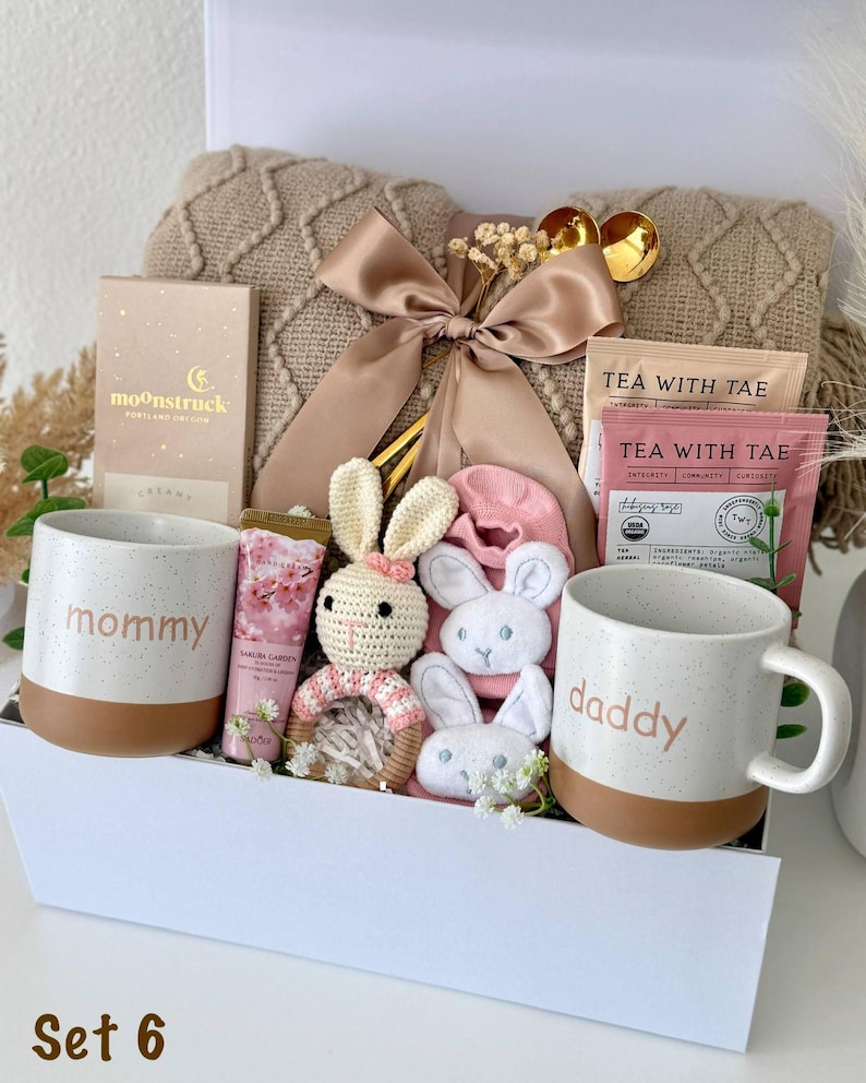 May include: A gift set containing a beige cable-knit blanket with a satin ribbon, two speckled ceramic mugs labeled mommy and daddy, a Moonstruck chocolate bar, Tea With Tae packets, a crochet bunny rattle, and pink baby socks in a white box.