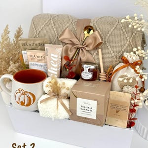 May include: A gift set in a white box containing a beige knit blanket with a satin bow, a pumpkin-print mug, tea packets, shea butter, a honey jar with a dipper, a gold spoon, a sea salt caramel box, oatmeal soap, and a small drawstring bag.