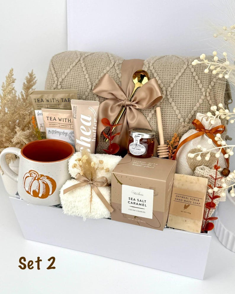 May include: A gift set in a white box containing a beige knit blanket with a satin bow, a pumpkin-print mug, tea packets, shea butter, a honey jar with a dipper, a gold spoon, a sea salt caramel box, oatmeal soap, and a small drawstring bag.