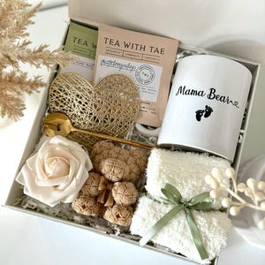 K&ouml;nnte beinhalten: Geschenkbox mit einer wei&szlig;en Tasse mit "Mama Bear" und Fu&szlig;abdr&uuml;cken, Teebeuteln, einem goldenen Herz, einem goldenen L&ouml;ffel, einer wei&szlig;en Rose, Teddyb&auml;r-Seifen und einem wei&szlig;en Waschlappen mit gr&uuml;nem Band. Die Box ist mit "Set 10" beschriftet.