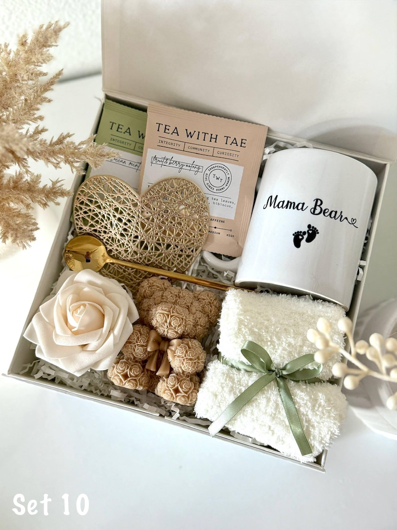 K&ouml;nnte beinhalten: Geschenkbox mit einer wei&szlig;en Tasse mit "Mama Bear" und Fu&szlig;abdr&uuml;cken, Teebeuteln, einem goldenen Herz, einem goldenen L&ouml;ffel, einer wei&szlig;en Rose, Teddyb&auml;r-Seifen und einem wei&szlig;en Waschlappen mit gr&uuml;nem Band. Die Box ist mit "Set 10" beschriftet.