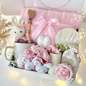 K&ouml;nnte beinhalten: Ein Baby-Geschenkset in einer wei&szlig;en Box mit einer rosa Decke, einem H&auml;schen aus H&auml;kelarbeit, einem herzf&ouml;rmigen Schnullerclip aus Holz und anderen Babyartikeln. Die Box tr&auml;gt den Text "CUTE BOXES Boutique" und "Set 12".