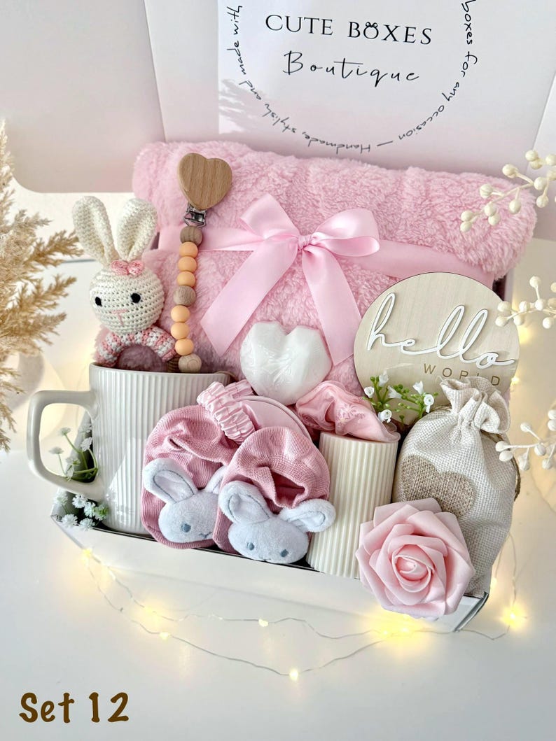 K&ouml;nnte beinhalten: Ein Baby-Geschenkset in einer wei&szlig;en Box mit einer rosa Decke, einem H&auml;schen aus H&auml;kelarbeit, einem herzf&ouml;rmigen Schnullerclip aus Holz und anderen Babyartikeln. Die Box tr&auml;gt den Text "CUTE BOXES Boutique" und "Set 12".