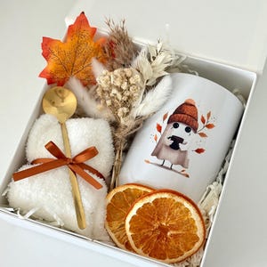 May include: A white gift box containing a white mug with a ghost illustration wearing a beanie, a pair of white fuzzy socks tied with a brown ribbon, a gold spoon, a dried floral bouquet, an artificial orange maple leaf, and two dried orange slices.