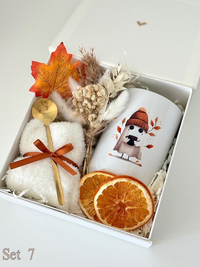 May include: A white gift box containing a white mug with a ghost illustration wearing a beanie, a pair of white fuzzy socks tied with a brown ribbon, a gold spoon, a dried floral bouquet, an artificial orange maple leaf, and two dried orange slices.