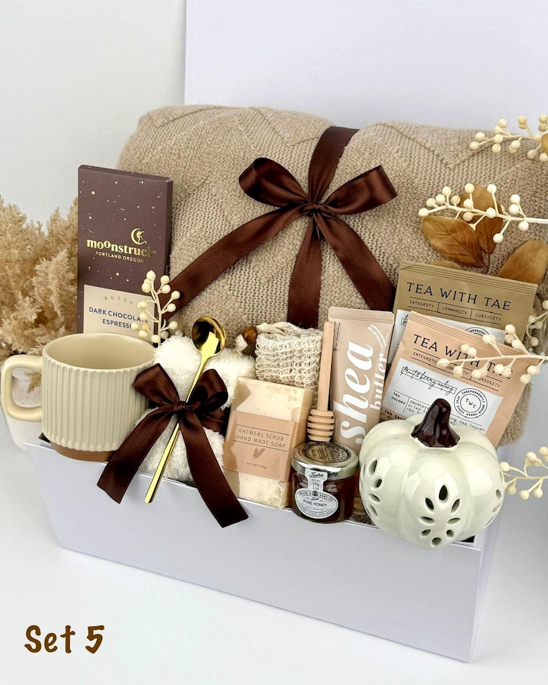 May include: A gift set in a white box containing a beige knit blanket with a brown ribbon, a Moonstruck dark chocolate bar, a ribbed beige mug, a gold spoon, oatmeal soap, a honey dipper, a jar of honey, shea butter, and a white ceramic pumpkin.