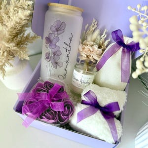May include: A lavender gift box contains a frosted glass tumbler with floral design and the name "Samantha", a small candle, a heart-shaped container of purple rose soaps, a small white towel, and a small bag, all tied with purple ribbons. The box is labeled "Set 12".