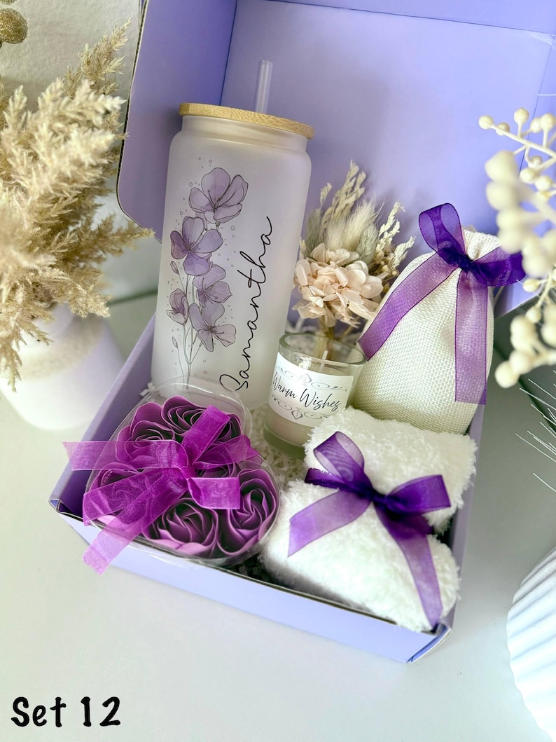 May include: A lavender gift box contains a frosted glass tumbler with floral design and the name "Samantha", a small candle, a heart-shaped container of purple rose soaps, a small white towel, and a small bag, all tied with purple ribbons. The box is labeled "Set 12".