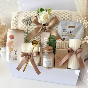 May include: A gift set containing a frosted glass tumbler with floral print and the name Beth, a cream-colored knit blanket, a Laikou snail collagen mask, a gold spoon, a small jar of bath salts, a bar of oatmeal soap, and a ribbed pillar candle.