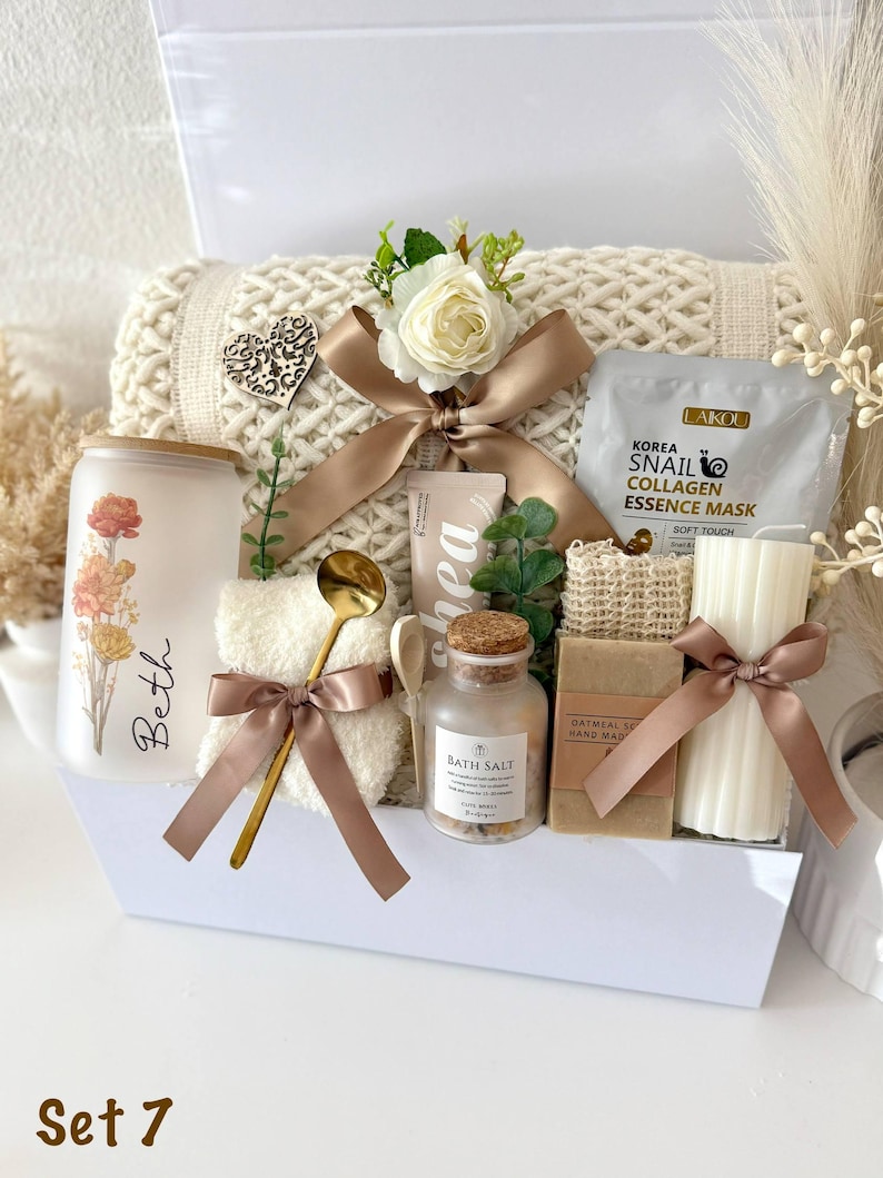 May include: A gift set containing a frosted glass tumbler with floral print and the name Beth, a cream-colored knit blanket, a Laikou snail collagen mask, a gold spoon, a small jar of bath salts, a bar of oatmeal soap, and a ribbed pillar candle.