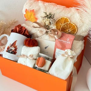 May include: A gift box containing a white mug with a gnome graphic, a cream-colored plush blanket tied with a ribbon, dried orange slices, a small pumpkin, a Moonstruck chocolate bar, a white drawstring pouch, and a candle with a pumpkin label.