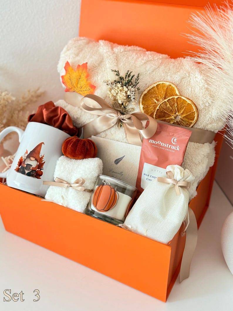 May include: A gift box containing a white mug with a gnome graphic, a cream-colored plush blanket tied with a ribbon, dried orange slices, a small pumpkin, a Moonstruck chocolate bar, a white drawstring pouch, and a candle with a pumpkin label.