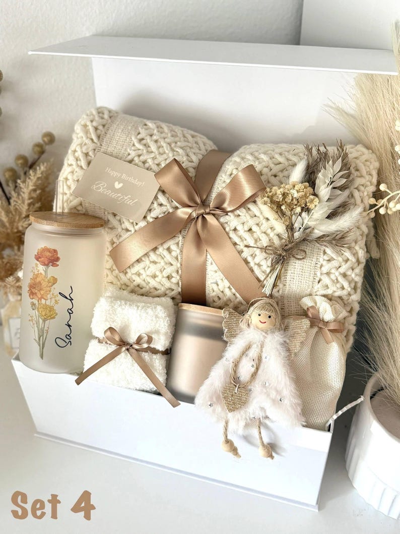 May include: A gift box set containing a cream-colored knitted blanket, a candle with floral design, a small towel, a decorative angel, and a small bag. The box is tied with a gold ribbon and includes a card that says "Happy Birthday! Beautiful."
