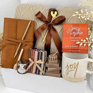 May include: A gift box containing a tan knit blanket with a brown ribbon and gold spoon, a brown leather journal with a gold pen, a jar of honey with a dipper, a patterned fabric roll, stacked book-shaped candles, and a mug with the word joy.