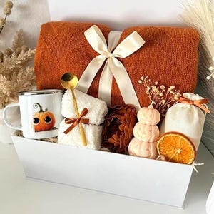 May include: A white gift box containing a folded orange knit blanket tied with a cream ribbon, a white mug with a pumpkin illustration, a gold spoon, a white fuzzy cloth, a stack of three pumpkin-shaped candles, and a dried orange slice.