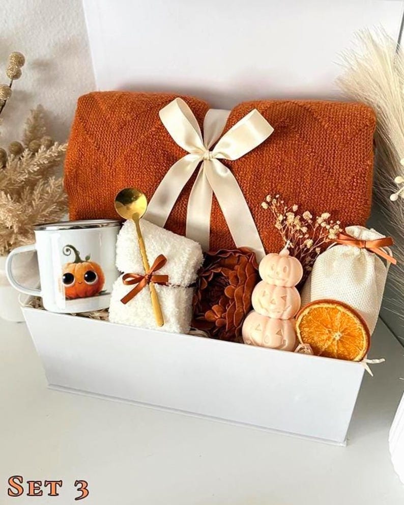 May include: A white gift box containing a folded orange knit blanket tied with a cream ribbon, a white mug with a pumpkin illustration, a gold spoon, a white fuzzy cloth, a stack of three pumpkin-shaped candles, and a dried orange slice.