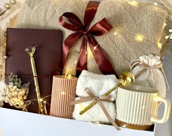 May include: A white gift box containing a beige knit blanket tied with a dark red ribbon, a dark brown notebook, a gold pen with a diamond-shaped topper, a ribbed peach candle, white fuzzy socks, a gold spoon, a mug, and a small drawstring pouch.