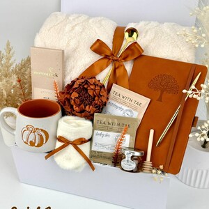 May include: A gift set containing a cream-colored fuzzy blanket tied with a rust-colored ribbon, a ceramic mug with a pumpkin graphic, a rust-colored notebook with a tree emblem, a gold pen, tea sachets, a small honey jar, and a wooden dipper.