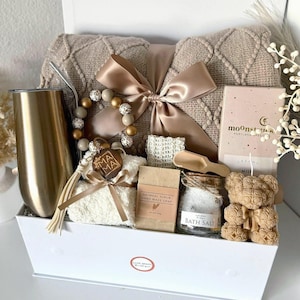 May include: A gift box containing a beige knit blanket with a satin ribbon, a gold tumbler, a beaded keychain with a wooden Mama tag, a bar of oatmeal scrub soap, a jar of bath salts with a wooden scoop, and a bear-shaped candle.