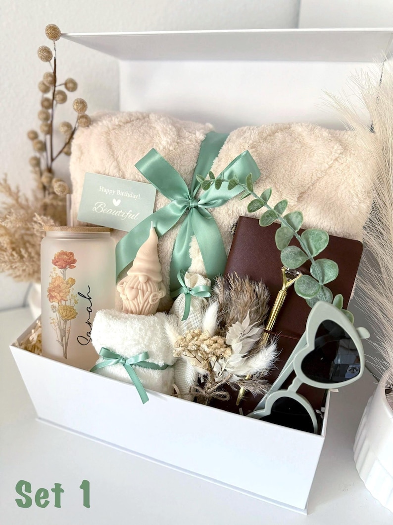 May include: A white gift box containing a beige faux-fur blanket tied with a sage green ribbon, a frosted glass tumbler with floral graphics and the name Sarah, a gnome figurine, a brown notebook, a gold pen, heart-shaped sunglasses, and dried flowers.