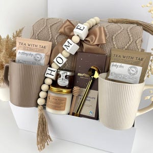 May include: A gift set containing two ribbed ceramic mugs, two Tea With Tae tea packets, a jar of honey, a candle, a Moons chocolate bar, a gold spoon, and a wooden honey dipper. A beige knit blanket and a wooden bead garland spelling HOME are included.