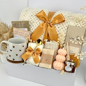 May include: A gift set containing a white knit blanket with a gold ribbon, a mug with brown hearts, two Tea With Tae packets, a gold spoon, a white washcloth, oatmeal soap, a pumpkin candle, a honey dipper, a jar of honey, and a chocolate bar.