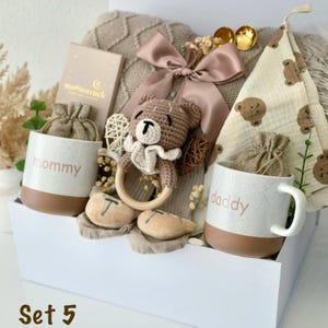 May include: A gift set in a white box containing two speckled ceramic mugs labeled mommy and daddy, a brown crocheted bear rattle with a wooden ring, a beige knit blanket with a satin bow, a bear-patterned cloth, and two small drawstring pouches.