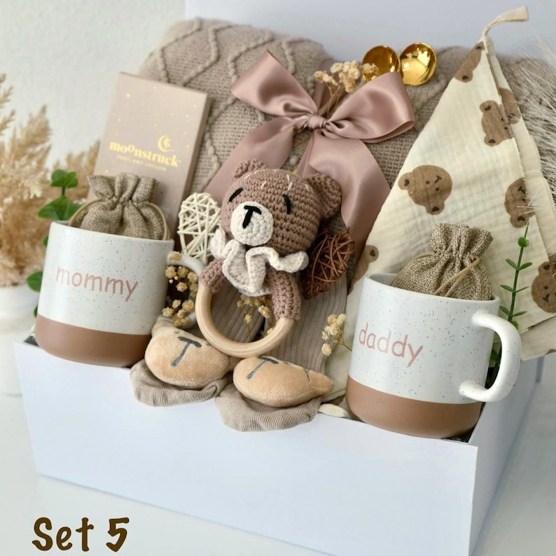 May include: A gift set in a white box containing two speckled ceramic mugs labeled mommy and daddy, a brown crocheted bear rattle with a wooden ring, a beige knit blanket with a satin bow, a bear-patterned cloth, and two small drawstring pouches.