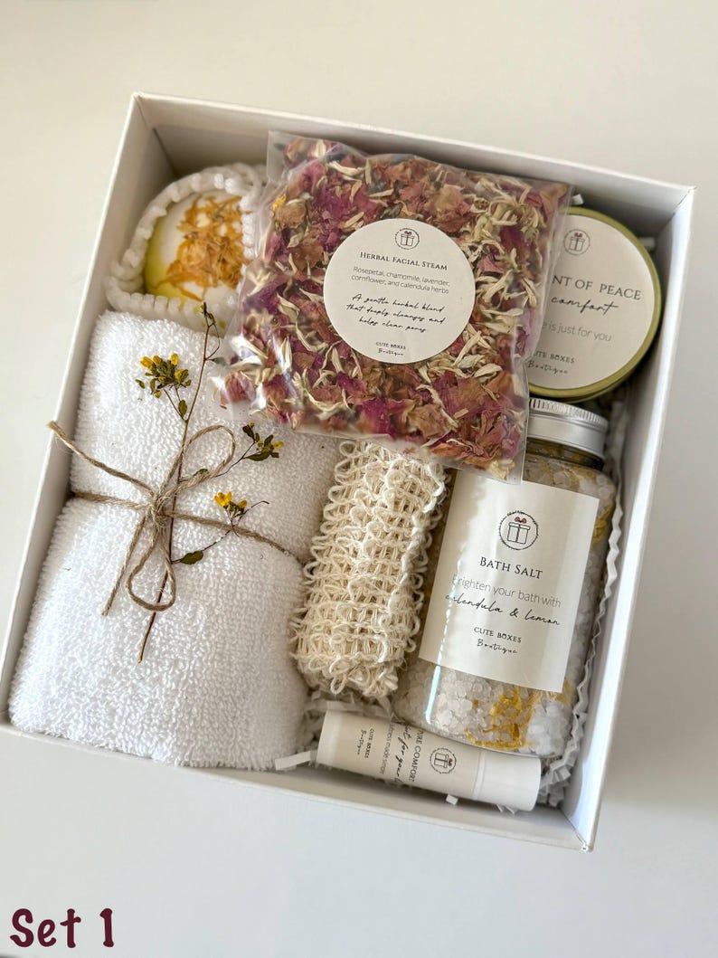 May include: A white gift box labeled "Set 1" filled with spa products. Includes a white washcloth, bath bomb, herbal facial steam, bath salt, and a lip balm. The box is tied with twine and decorated with dried flowers.