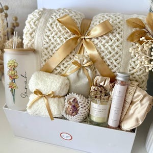 May include: A gift set in a white box, featuring a cream-coloured knitted blanket tied with a gold ribbon. Includes a glass tumbler with floral design, a candle, bath salts, a sleep mask, and other items.
