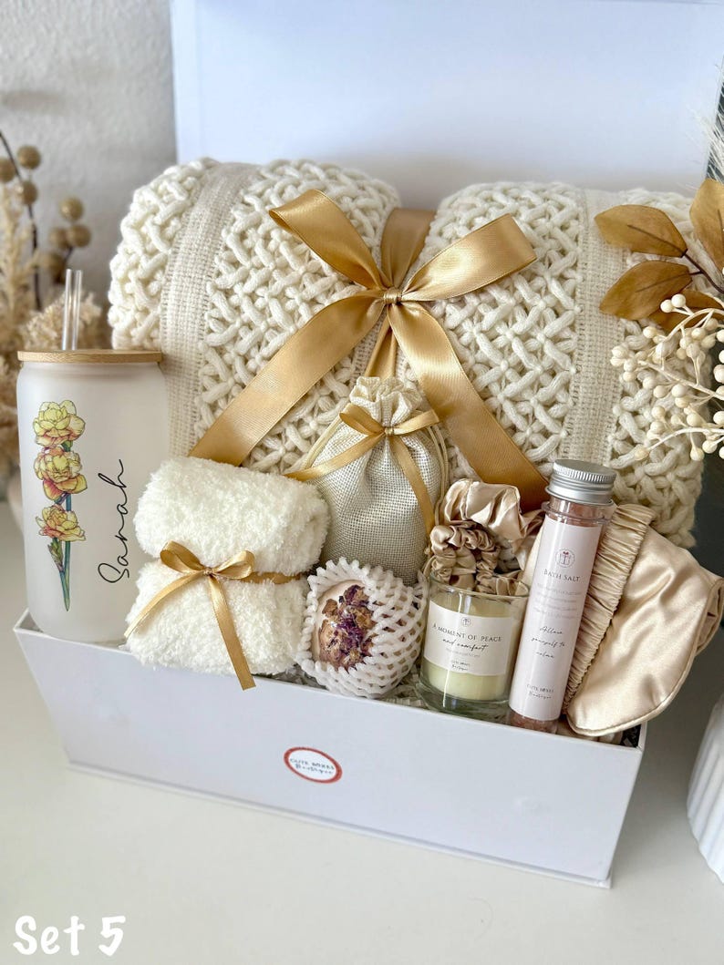 May include: A gift set in a white box, featuring a cream-coloured knitted blanket tied with a gold ribbon. Includes a glass tumbler with floral design, a candle, bath salts, a sleep mask, and other items.
