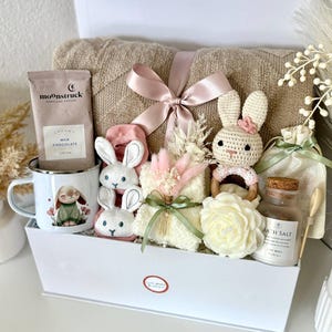 K&ouml;nnte beinhalten: Eine wei&szlig;e Geschenkbox gef&uuml;llt mit einer beigen Decke, einer Tasse mit Hasenillustration, einer Kakaopackung, Pl&uuml;sch-Hasenspielzeug, einem geh&auml;kelten Hasenrassel, Badesalz und einer dekorativen Blume. Die Box ist mit einem rosa Band gebunden.
