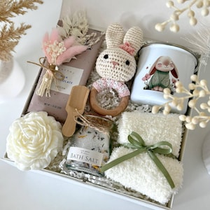 K&ouml;nnte beinhalten: Geschenkbox mit geh&auml;keltem Hasenrassel, Tasse mit Hasenmotiv, Schokoriegel, Badesalz, Kerze und flauschigem wei&szlig;en Handtuch mit gr&uuml;nem Band. Die Box ist mit "Set 5" beschriftet. Die Artikel sind auf wei&szlig;em Papier angeordnet.