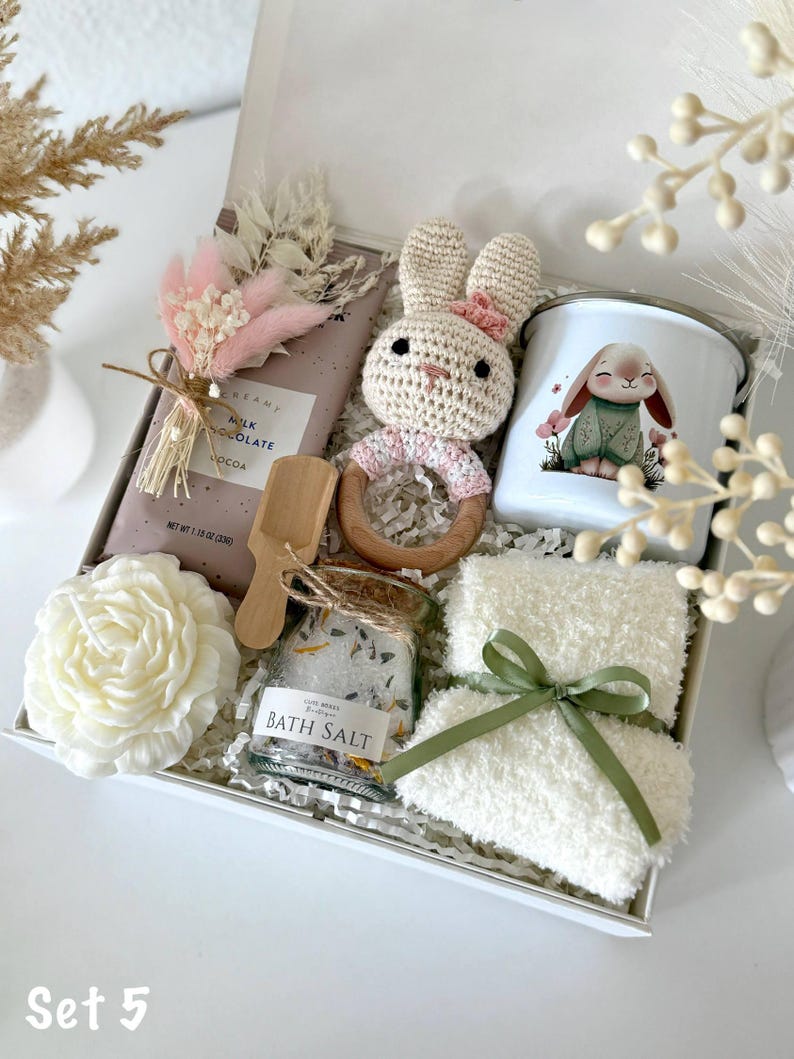 K&ouml;nnte beinhalten: Geschenkbox mit geh&auml;keltem Hasenrassel, Tasse mit Hasenmotiv, Schokoriegel, Badesalz, Kerze und flauschigem wei&szlig;en Handtuch mit gr&uuml;nem Band. Die Box ist mit "Set 5" beschriftet. Die Artikel sind auf wei&szlig;em Papier angeordnet.