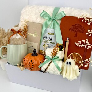 May include: A gift set in a white box containing a ribbed light green mug, a Moonstruck milk chocolate bar, a Laikou snail collagen essence mask, a brown notebook with a tree design, a bamboo pen, a pumpkin-shaped item, and a ghost-shaped candle.