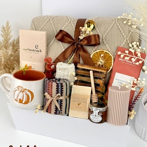 May include: A gift set containing a beige knit blanket with a brown ribbon, a white mug with an orange pumpkin, two Moonstruck chocolate bars, a patterned cloth, oatmeal soap, a wooden honey dipper, a jar of honey, and a ribbed candle.