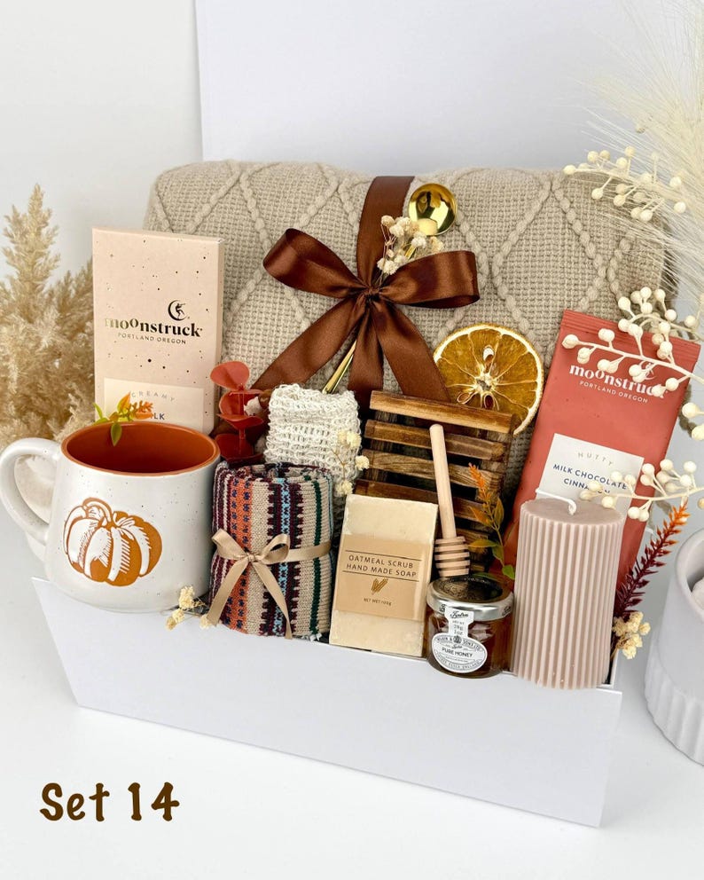 May include: A gift set containing a beige knit blanket with a brown ribbon, a white mug with an orange pumpkin, two Moonstruck chocolate bars, a patterned cloth, oatmeal soap, a wooden honey dipper, a jar of honey, and a ribbed candle.
