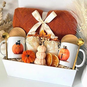 May include: A white gift box containing two white ceramic mugs with pumpkin illustrations, two gold-toned spoons, two wooden lids, a folded rust-colored knit blanket tied with a cream ribbon, two small fabric pouches, and decorative dried fruit.
