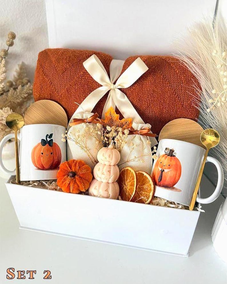 May include: A white gift box containing two white ceramic mugs with pumpkin illustrations, two gold-toned spoons, two wooden lids, a folded rust-colored knit blanket tied with a cream ribbon, two small fabric pouches, and decorative dried fruit.