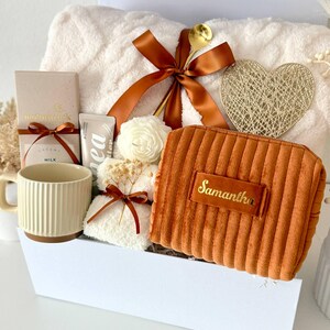 May include: A gift basket filled with self-care items. It includes a plush white towel, a ribbed mug, a small orange cosmetic bag with the name "Samantha", a gold spoon, and a decorative heart.