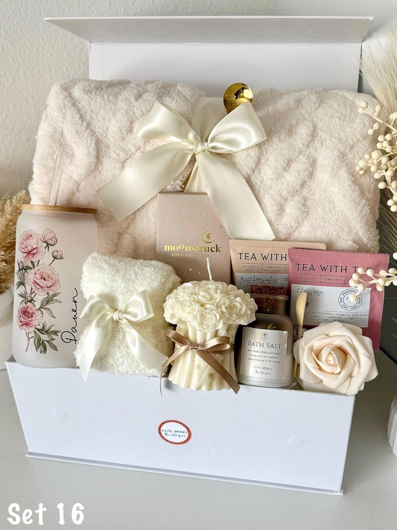 May include: A white gift box filled with self-care items. Includes a fluffy blanket, a floral-decorated glass, a small towel with a bow, a candle, bath salts, tea, and a rose-shaped soap. The box is tied with a large white bow.