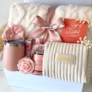 May include: A gift set containing a cream-colored textured throw blanket tied with a dusty pink ribbon, a matte pink tumbler with a straw, a pink scrunchie, a small candle, a rose, a pouch labeled Sarah, and a red tea box labeled Cup of Love.