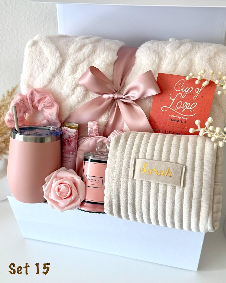 May include: A gift set containing a cream-colored textured throw blanket tied with a dusty pink ribbon, a matte pink tumbler with a straw, a pink scrunchie, a small candle, a rose, a pouch labeled Sarah, and a red tea box labeled Cup of Love.