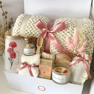 May include: A white gift box containing self-care products. It features a cream-coloured knitted blanket, a glass with a floral design, bath salts, soap, a candle, and a small bouquet, all tied with pink ribbons.