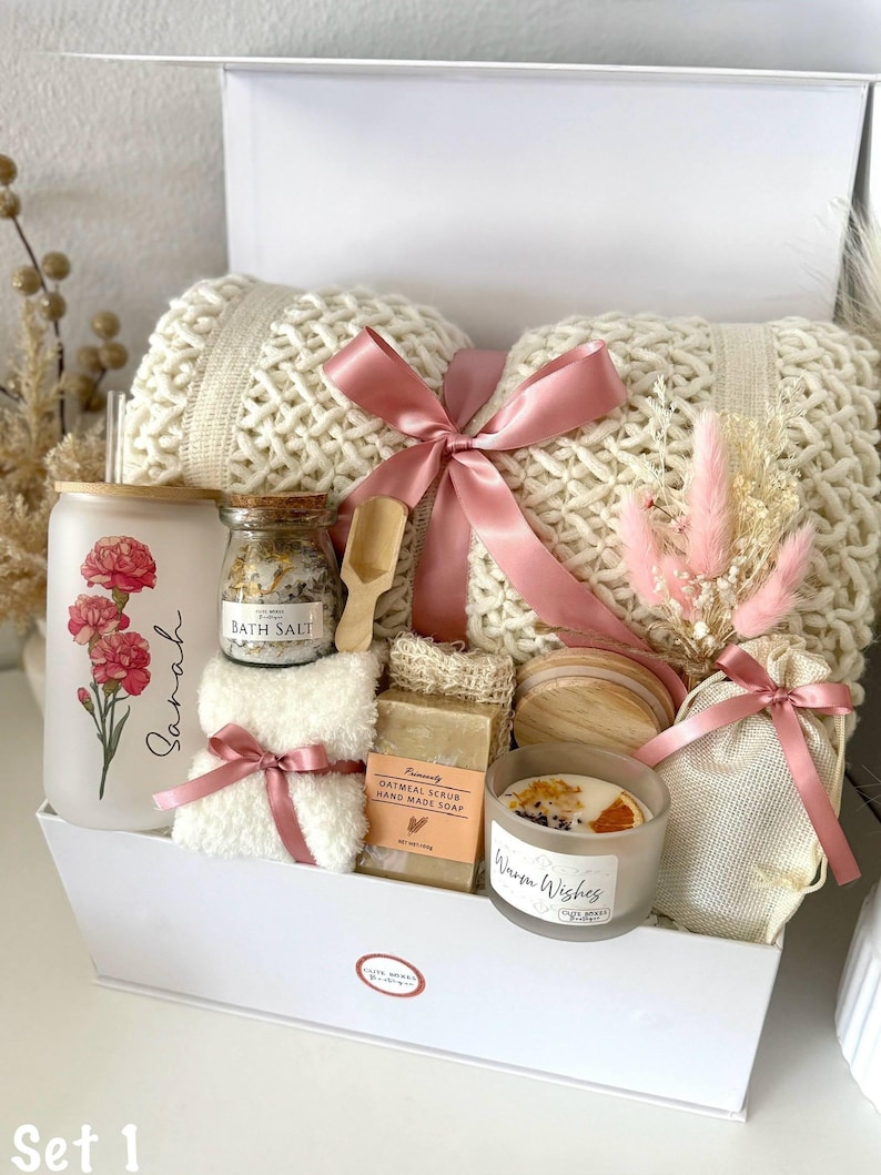 May include: A white gift box containing self-care products. It features a cream-coloured knitted blanket, a glass with a floral design, bath salts, soap, a candle, and a small bouquet, all tied with pink ribbons.