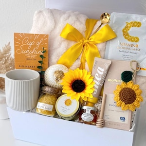 May include: A white gift box containing a cream-colored plush blanket with a yellow ribbon, a ribbed mug, a box of Cup of Sunshine tea, a Vitamin C mask, a jar of yellow bath salts, a Hug in a Jar candle, a sunflower, shea butter, honey, and a chocolate bar.