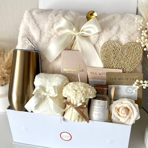 Puede incluir: Una caja de regalo blanca llena de art&iacute;culos de cuidado personal. Incluye una manta de felpa, un vaso dorado, una vela, sales de ba&ntilde;o, t&eacute; y un coraz&oacute;n decorativo. La caja est&aacute; atada con un lazo color crema. El texto "Set 6" est&aacute; abajo.