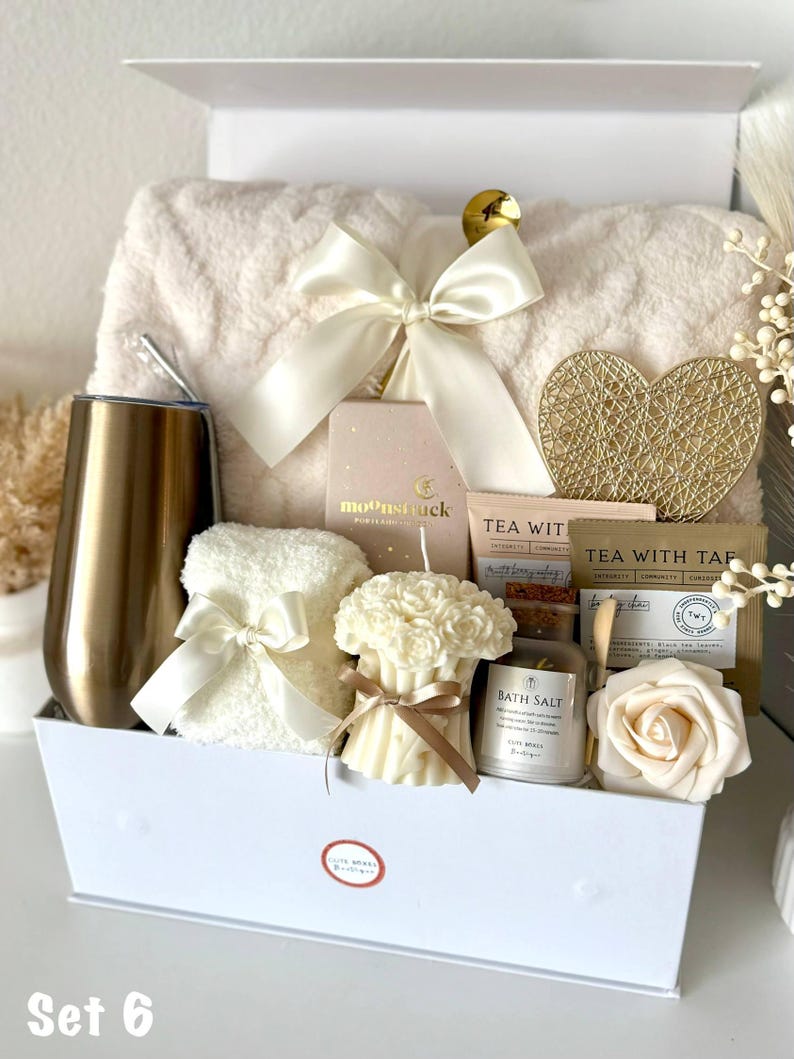 Puede incluir: Una caja de regalo blanca llena de art&iacute;culos de cuidado personal. Incluye una manta de felpa, un vaso dorado, una vela, sales de ba&ntilde;o, t&eacute; y un coraz&oacute;n decorativo. La caja est&aacute; atada con un lazo color crema. El texto "Set 6" est&aacute; abajo.