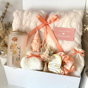 May include: A white gift box filled with a peach-colored towel, a glass tumbler with floral design and the name "Sarah", a gnome candle, scrunchies, and a small gift bag. A card reads "Happy Birthday! Beautiful".